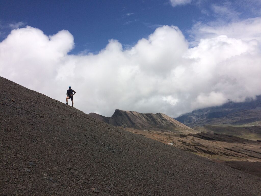 A high-altitude hiker stands on a steep, rocky slope beneath dramatic clouds and jagged peaks—capturing the science of joy as a compass: a moment of alignment between effort, meaning, and awe in the midst of challenge.