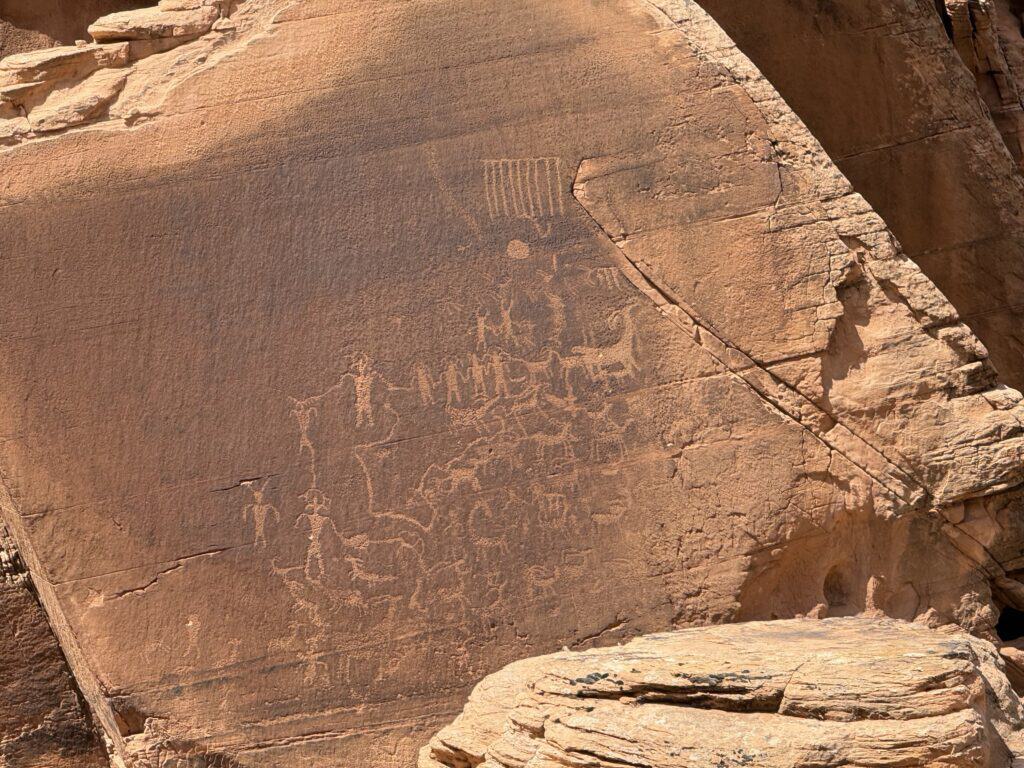Ancient petroglyphs pecked into a sunlit sandstone cliff in southeastern Utah, showing human figures, animals, and abstract markings on the desert rock face.