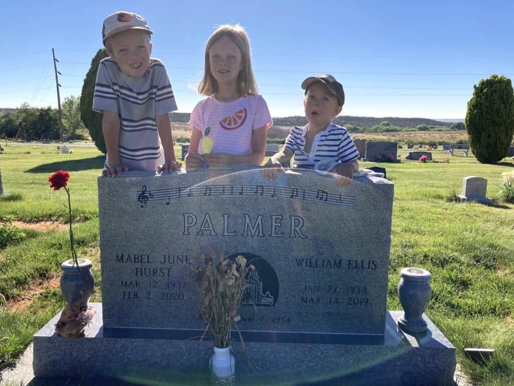 Three young children stand smiling behind a granite headstone in the Blanding, Utah cemetery. The stone reads PALMER and bears the names of Mabel June Hurst Palmer (1937-2020) and William Ellis Palmer (1934-2019), with a hand-engraved musical staff and notes carved across the top, the melody of "The Land I Love." Open grassland and juniper hills stretch behind them.

