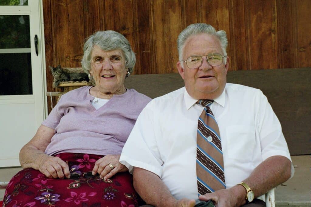 Mabel and William Ellis Palmer, the writer's maternal grandparents, sit together against a wood-paneled wall of John Bishoff's childhood home in Moab, Utah. She wears a lavender shirt and floral skirt; he wears a white short-sleeved shirt and a striped tie. A tabby cat is curled on a shelf behind them.