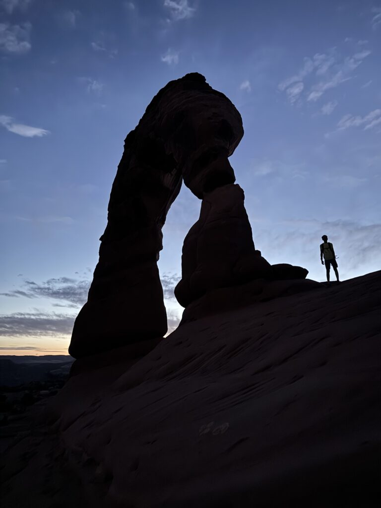 Lone silhouetted figure standing beside Delicate Arch at dusk in Moab, Utah, reflecting in the quiet.