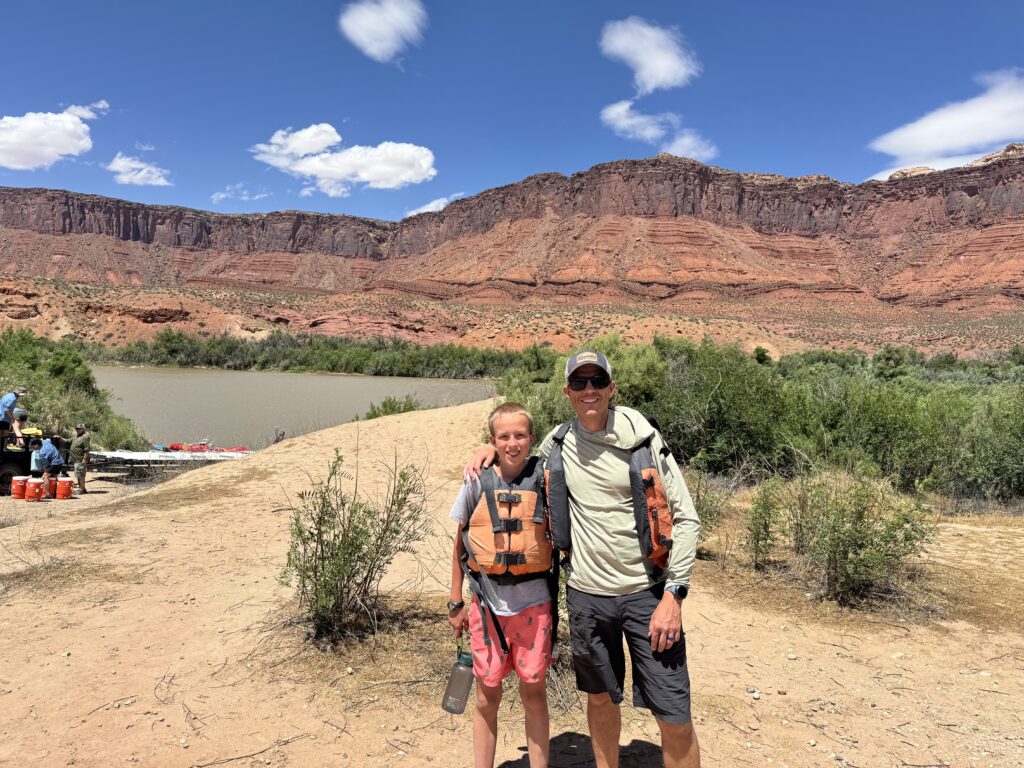 Father and son in life jackets smiling together on the Colorado River beneath red rock cliffs in Moab, Utah.